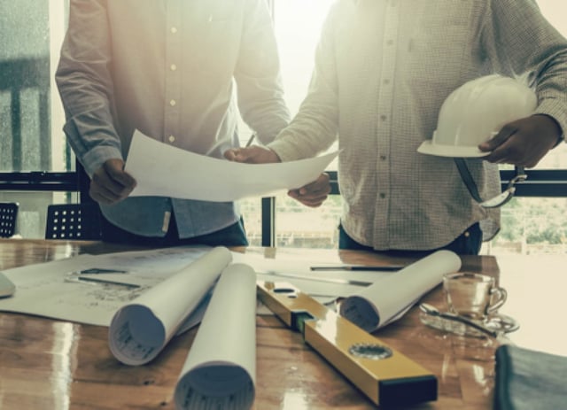 Two men discuss project plans in front of a wall of windows.