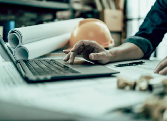 A person works on a laptop next to a stack of rolled-up plans.