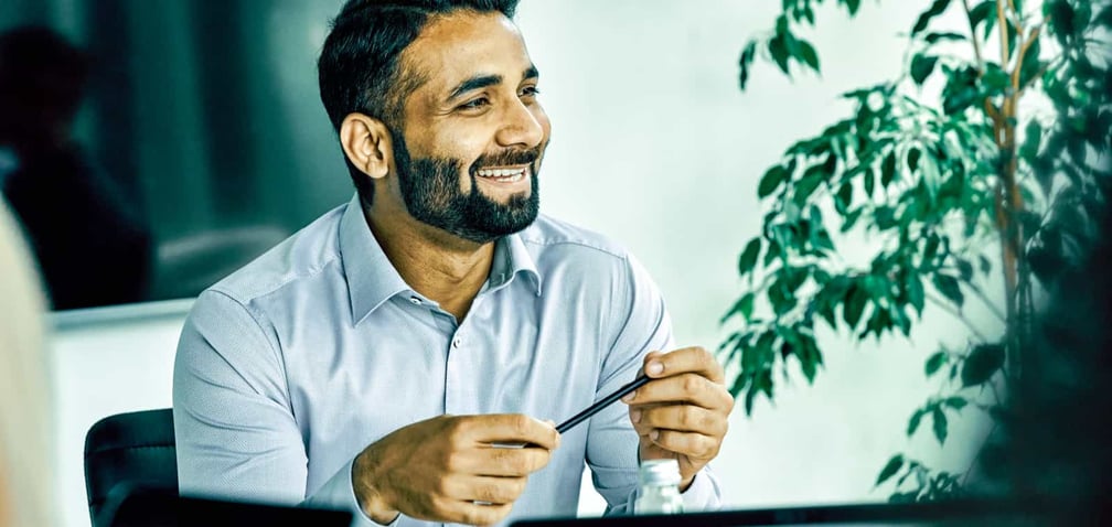 A smiling man in a blue shirt holds a pencil during a meeting in an office.