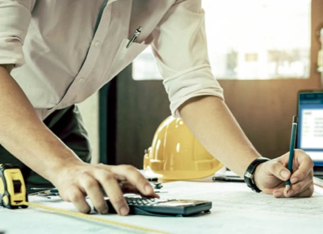 A person leans over a desk while working with a calculator and a pen.